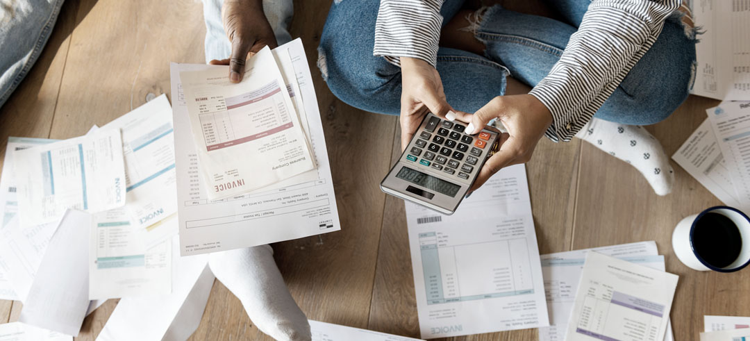 Dos personas sentadas en el suelo rodeadas de facturas, revisando y sumando cuentas