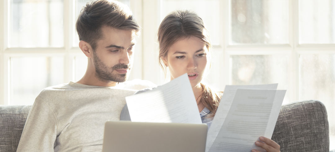 Pareja sentada en el sofá mientras revisan diferentes documentos físicos y en el ordenador
